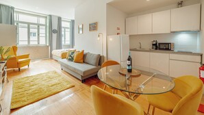 A modern living room with a sofa, yellow chairs, a glass table, and a kitchen area with cabinets and appliances. - Downtown Suites by Vacationy (Porto)
