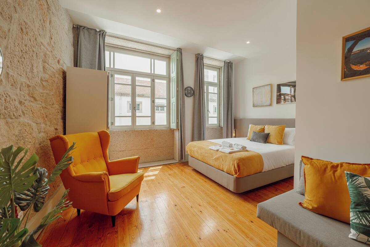 Une chambre d’hôtel moderne dotée d’un grand lit, d’un canapé, d’un fauteuil et offrant une vue sur la ville grâce à de grandes fenêtres.