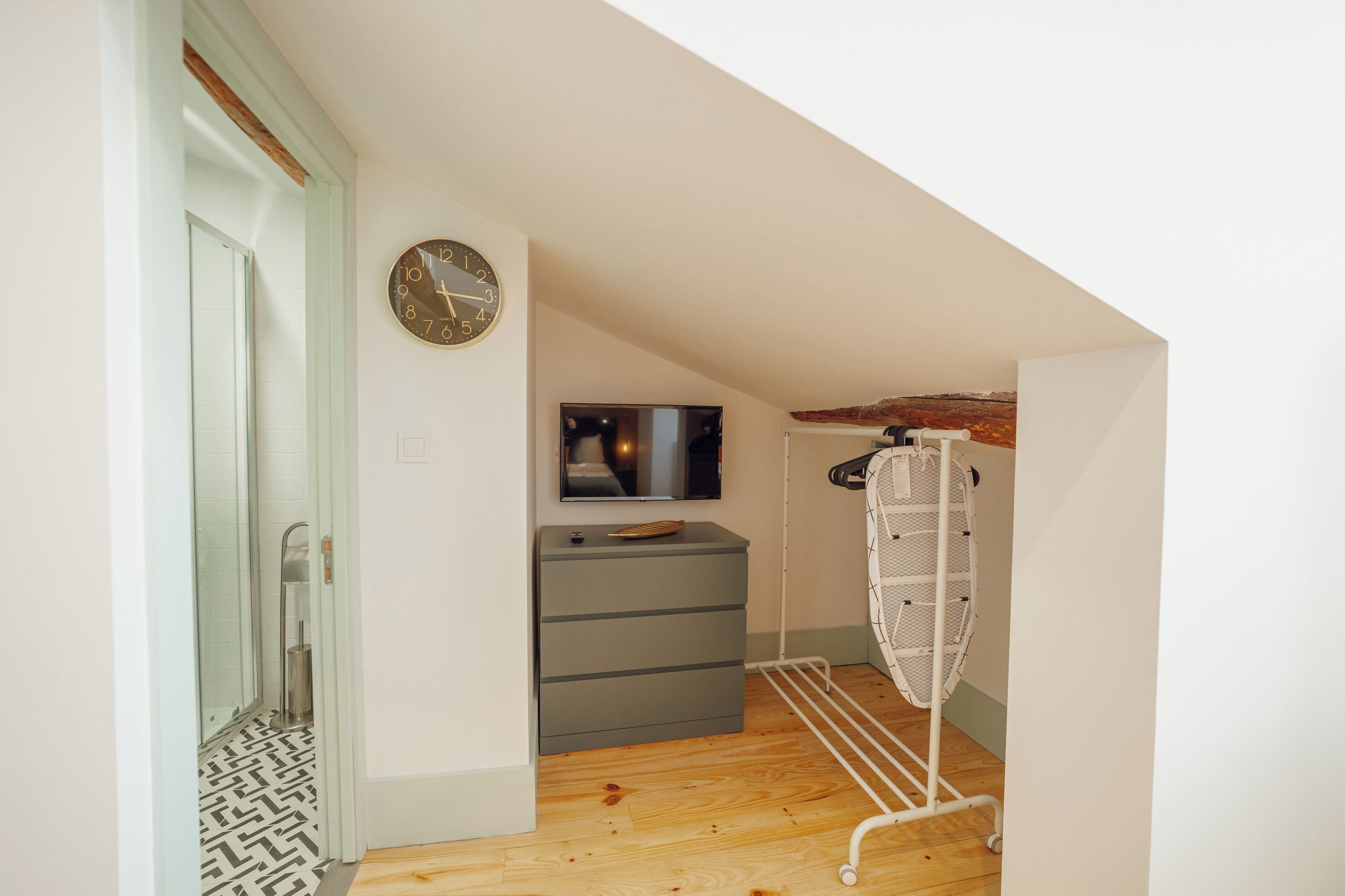 A small, well-lit room with a wooden floor, a grey dresser, a wall-mounted TV, and a drying rack.