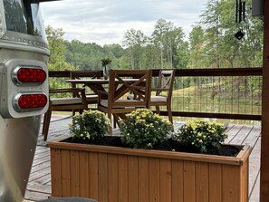 Eine Holzterrasse mit Tisch und Stühlen, ein Blumenkasten mit gelben Blüten und im Hintergrund ein See und Bäume.