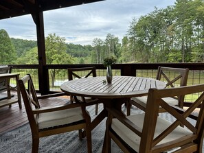 Eine Holzterrasse mit rundem Tisch und Stühlen, mit Blick auf einen See und einen bewaldeten Bereich.
