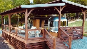 A wooden cabin with a covered patio, a grill, and a camper parked inside.