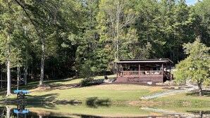 A wooden cabin with a covered deck, surrounded by a pond and dense forest.