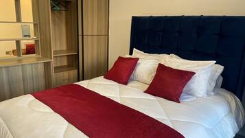 Una cama bien tendida con ropa de cama blanca y roja, un cabecero oscuro y un armario de madera con espejo y repisas.
