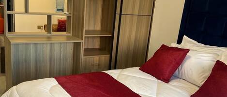 Una cama bien hecha con ropa de cama blanca y roja, un armario de madera con espejo y una pequeña zona de escritorio.