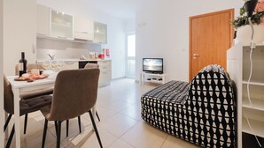 Un salon moderne avec un canapé, un coin repas comprenant une table et des chaises, une cuisine avec des meubles blancs et un téléviseur posé sur un meuble.