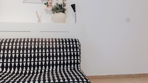 Une pièce avec un canapé à motifs, une étagère fixée au mur sur laquelle est posé un vase de fleurs, et un tableau.