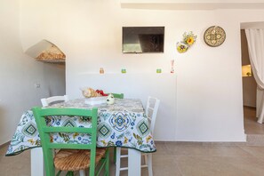Zona de menjador amb taula coberta per un mantell estampat, envoltada de cadires blanques amb detalls verds. Un televisor de pantalla plana està fixat a la paret sobre la taula.