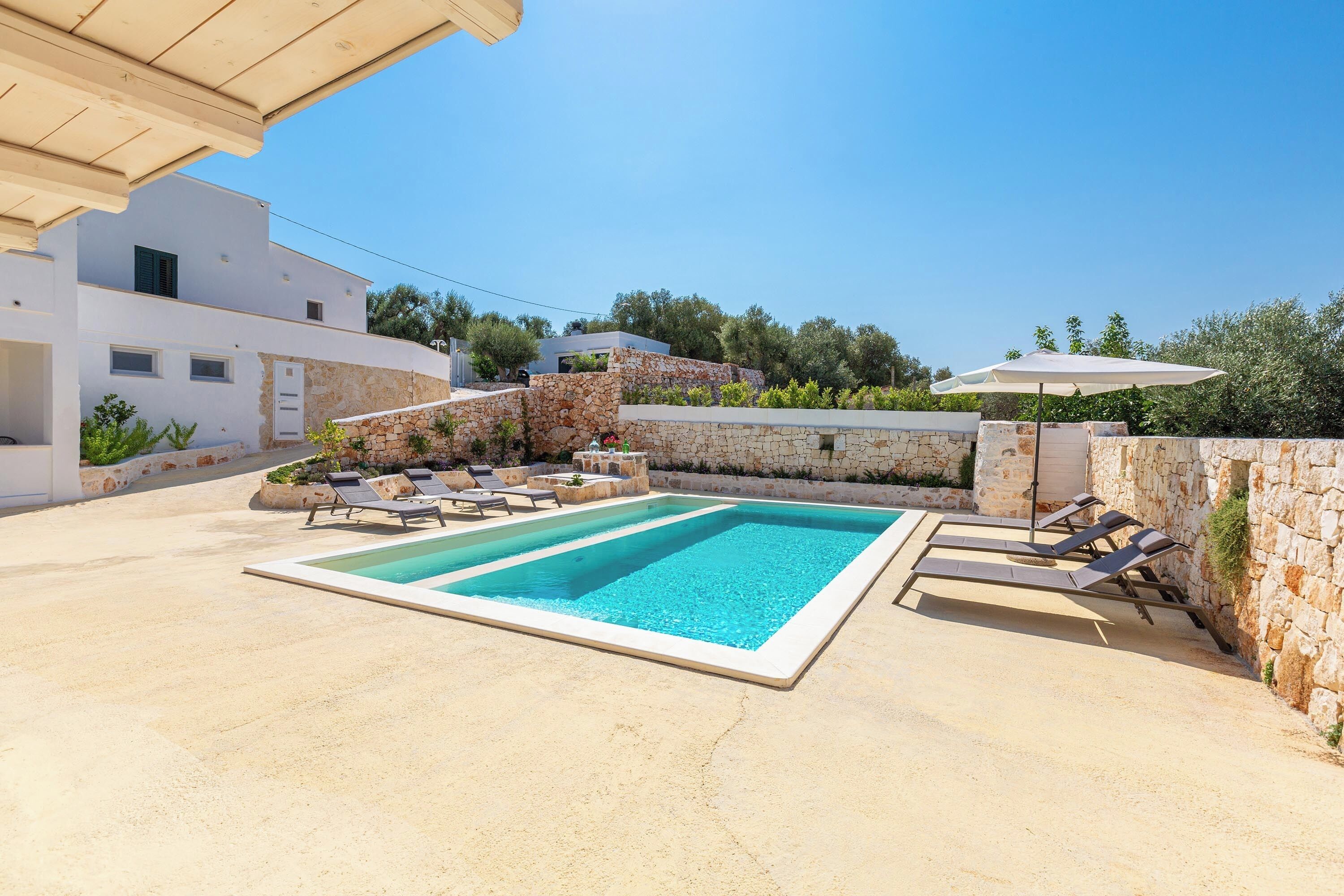 A rectangular swimming pool with lounge chairs and umbrellas, surrounded by stone walls and vegetation.