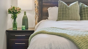 Une chambre avec un grand lit, une table de chevet avec une lampe et un vase de fleurs, une fenêtre avec des rideaux à motifs et une vue sur l’extérieur.