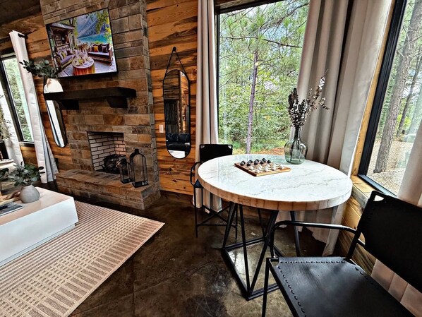 A cozy room with a stone fireplace, a round table with a chessboard, and a large window offering a view of trees.