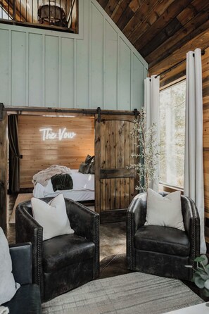A cozy room with a bed, two leather armchairs, a wooden door with a neon sign, and a large window.