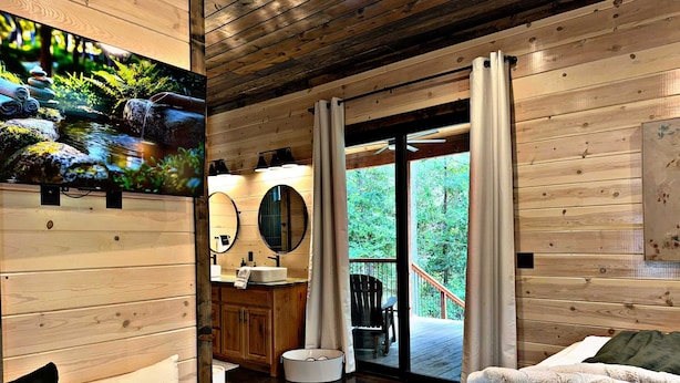 Interior de una cabaña de madera con baño, una cama y un balcón con vistas al bosque.
