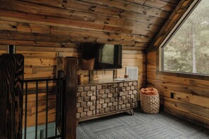 A wooden cabin interior with a TV mounted on the wall, a stone fireplace, and a window with a view of trees.