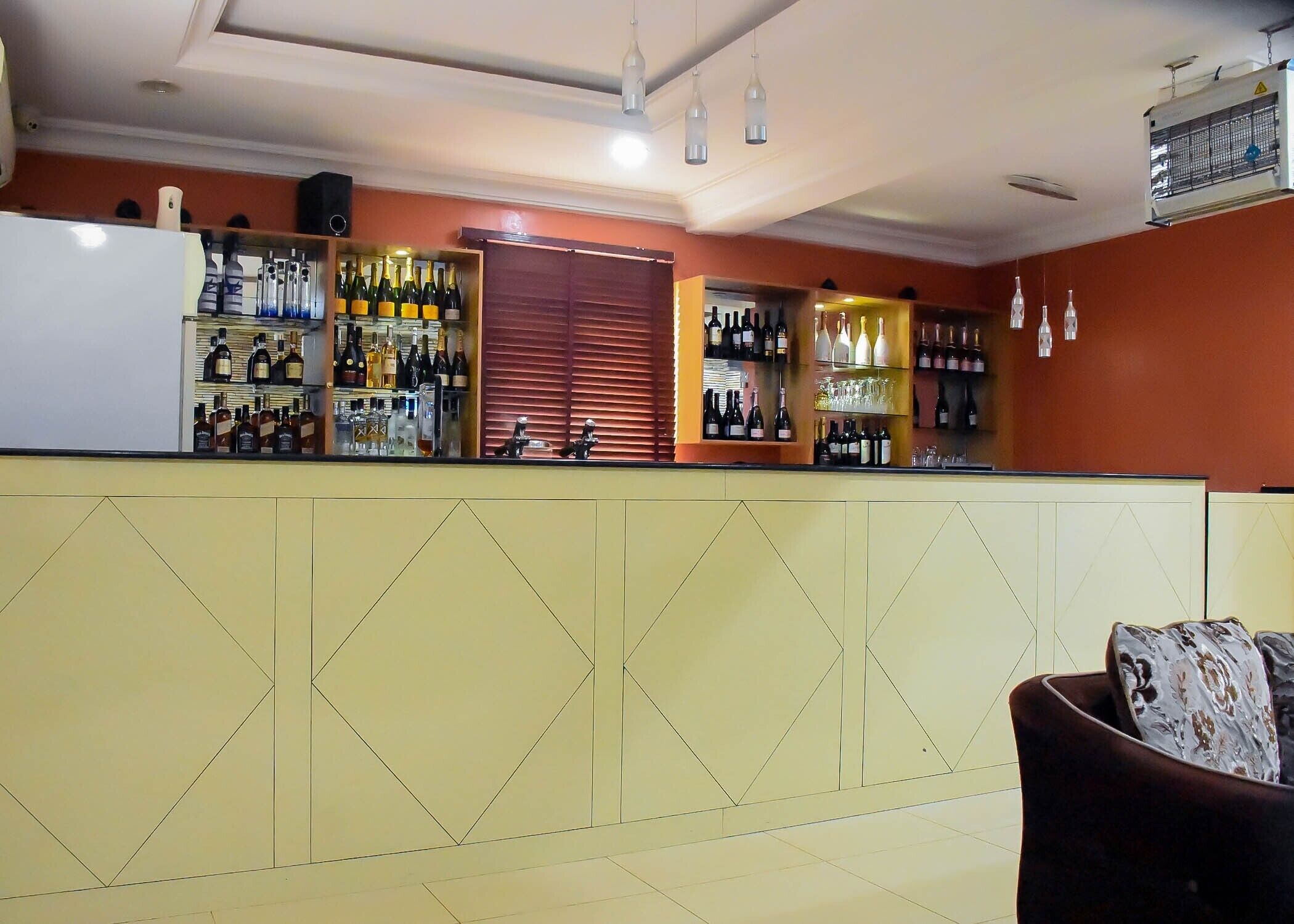 A bar area with shelves of bottles and glasses, a counter, and a seating area with a patterned cushion.