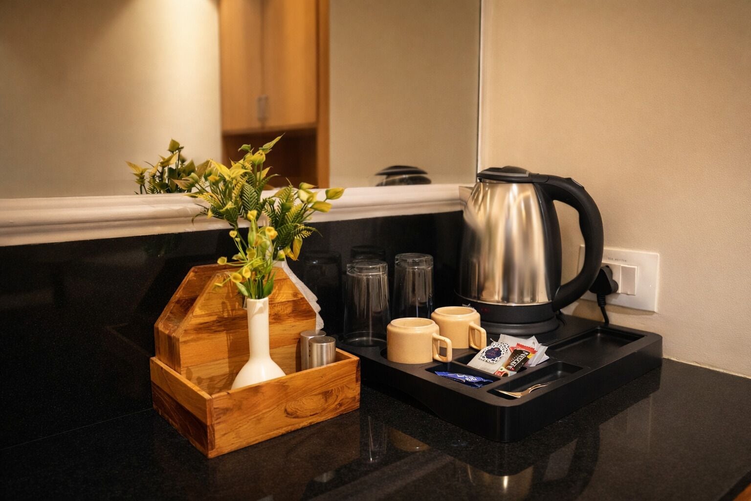 En köksbänk med en kaffebryggare, två muggar, en trälåda med en vas och en liten växt.