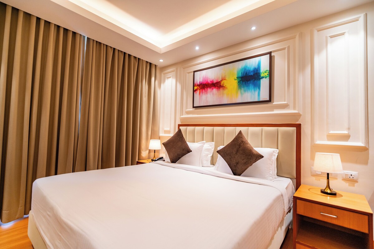 A hotel room with a large bed, a nightstand with a lamp, a painting on the wall, and a window with curtains.