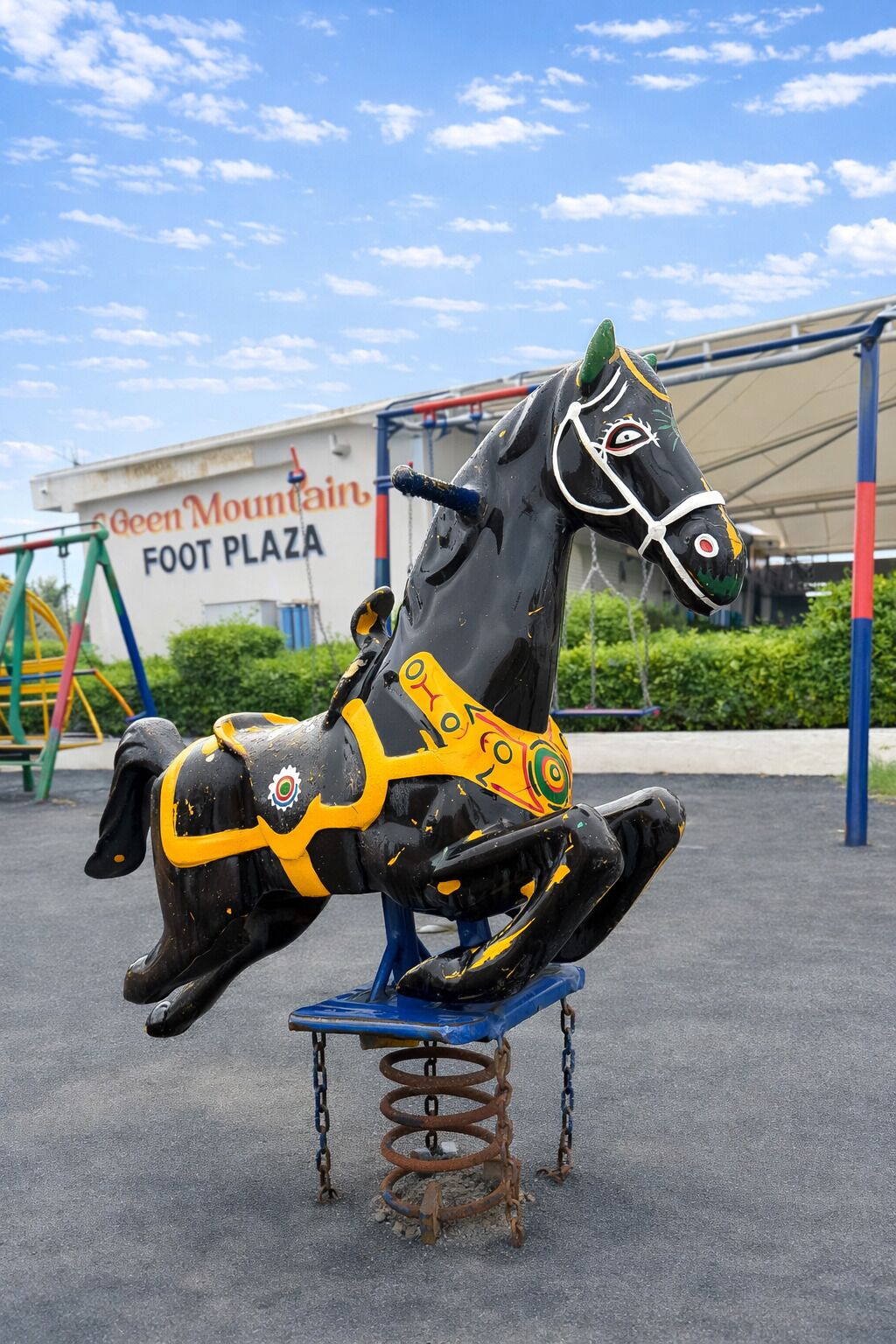 A black and yellow carousel horse at a playground with a sign reading "Green Mountain Foot Plaza" in the background.