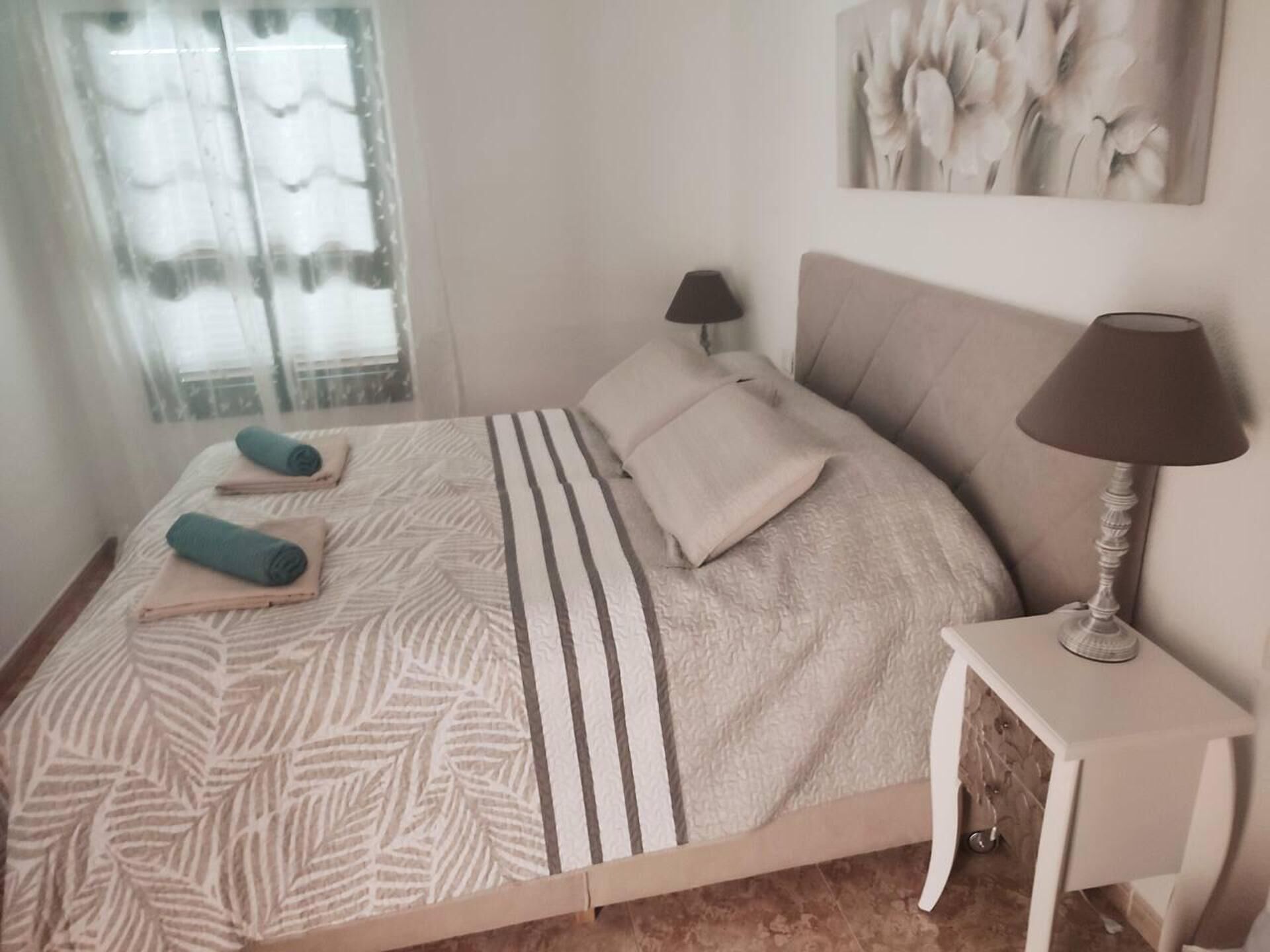 A neatly made bed with a striped blanket and two rolled towels, a bedside lamp, and a painting on the wall.