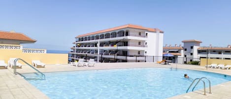 A pool area with a clear view of a multi-story building with balconies, a tiled deck, and lounge chairs.