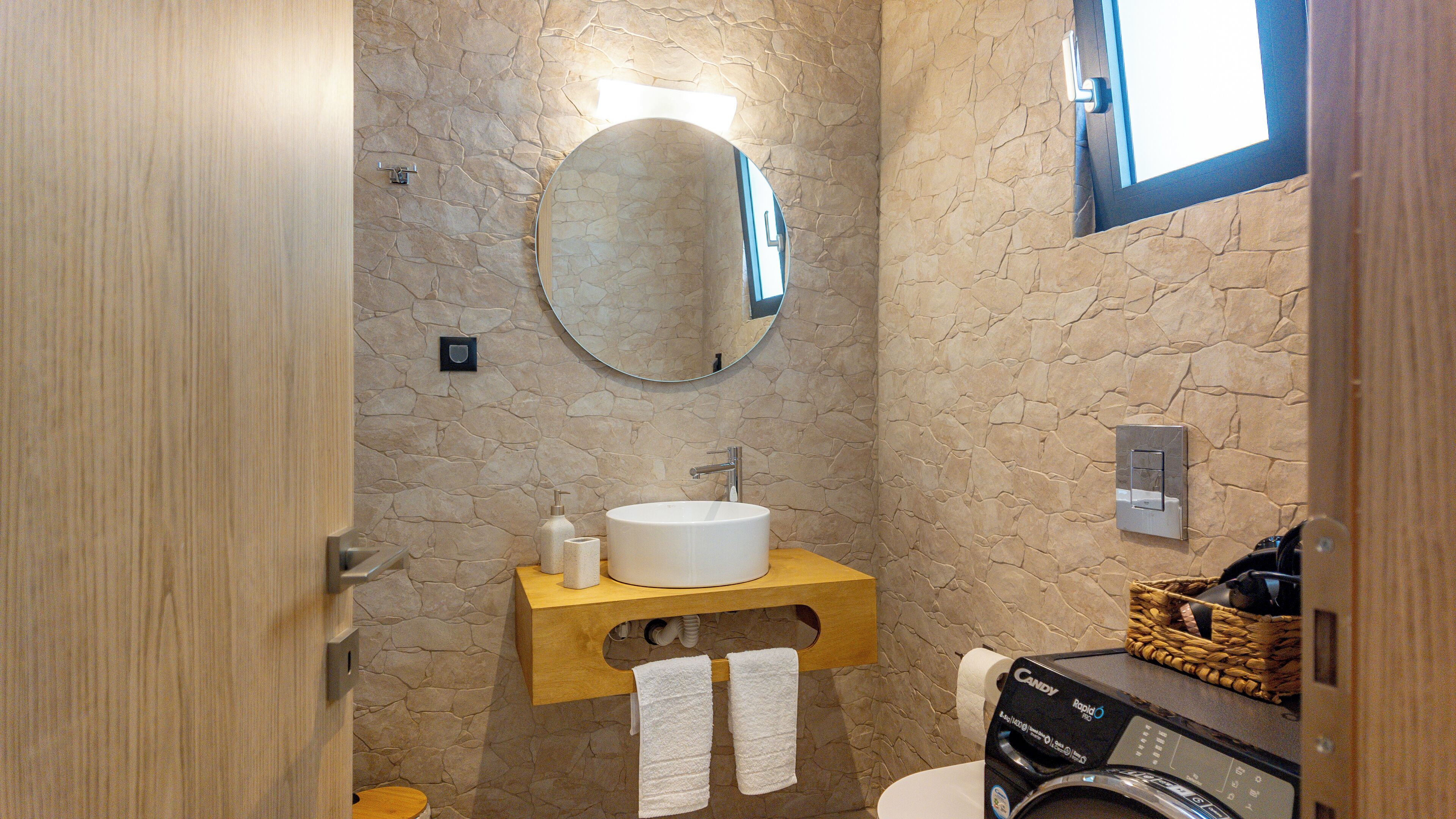 Une salle de bain avec un mur en pierre, un meuble-lavabo en bois, un miroir rond, un lavabo blanc et un lave-linge.