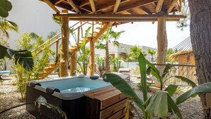 Un jacuzzi bajo una pérgola de madera, con una palmera y sillones de descanso al fondo.
