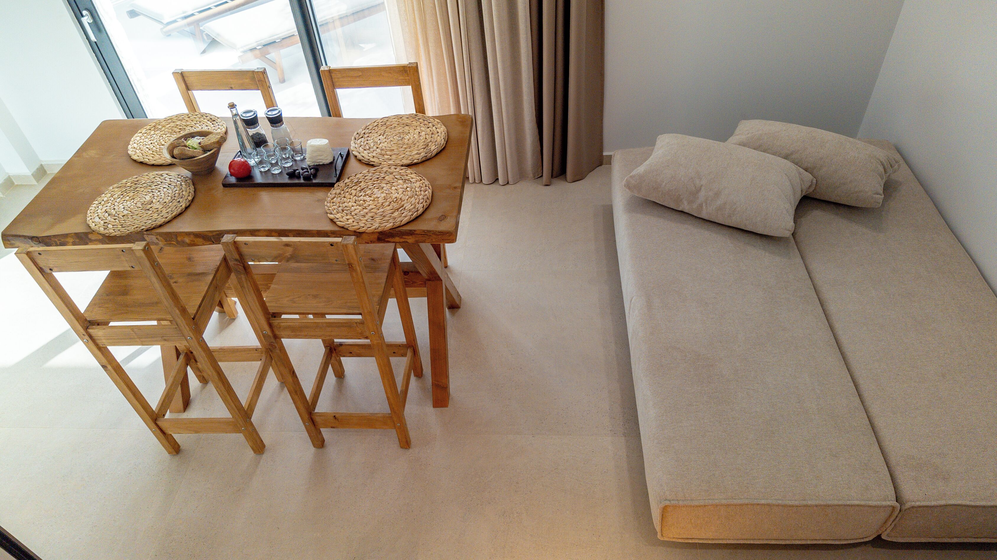 A wooden dining table with woven placemats, a bottle, and glasses. Beside the table is a beige sofa with cushions.