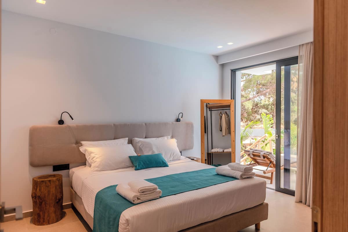 A modern bedroom with a large bed, a wooden nightstand, and a view of the outdoors through a sliding glass door.