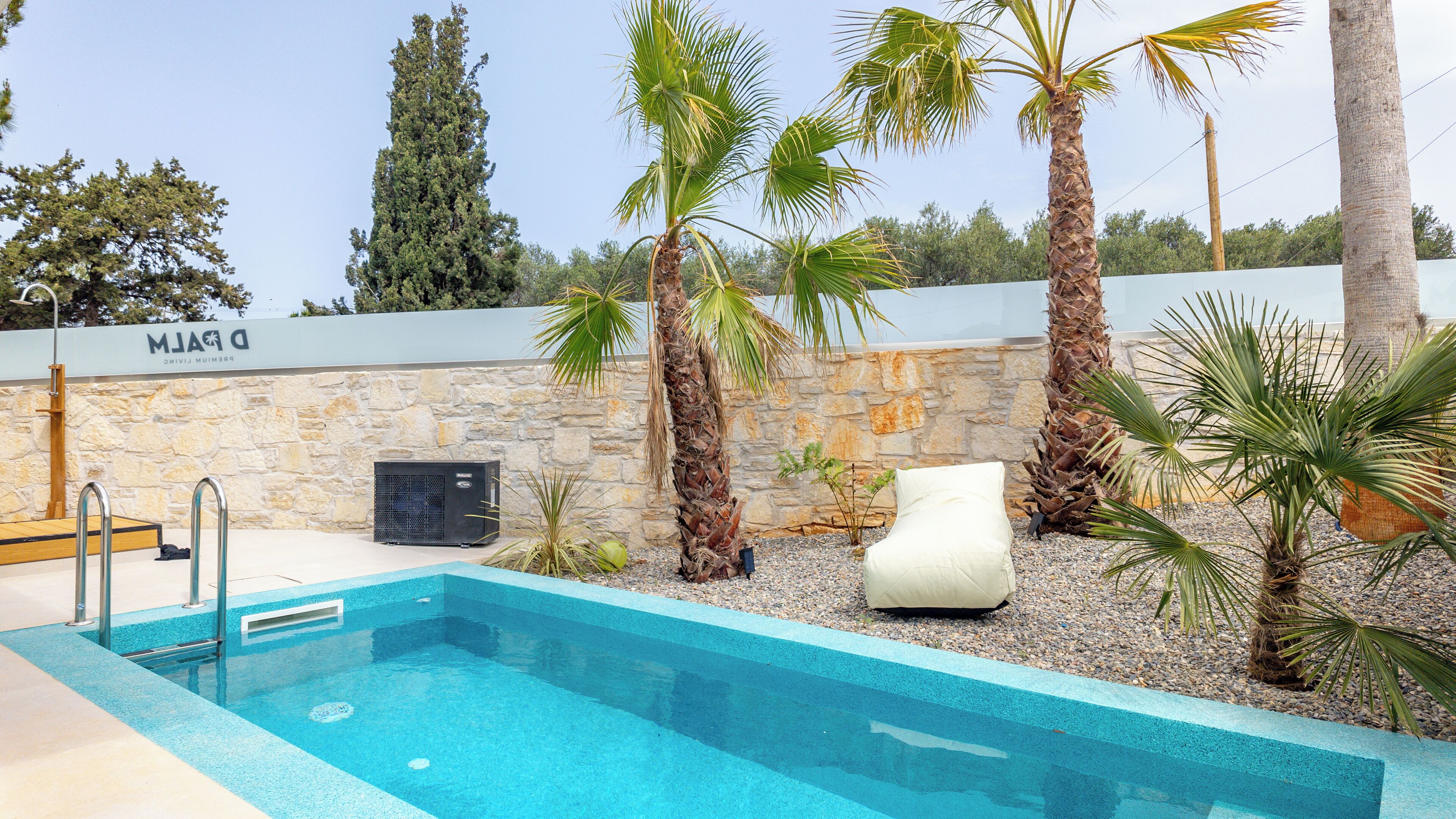 A swimming pool with a stone wall, palm trees, and a white lounge chair.