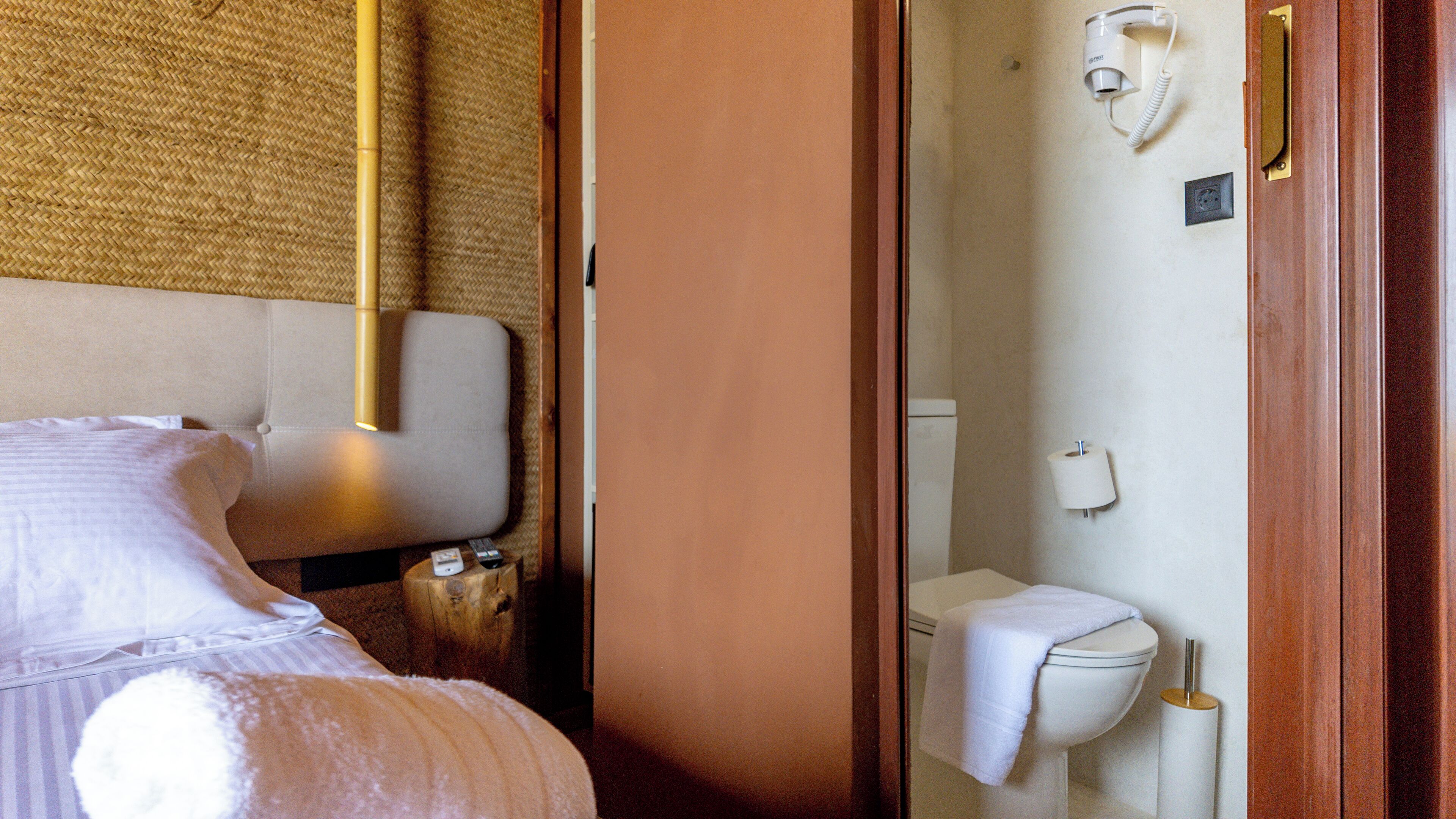 A hotel room with a bed, a bedside table, a lamp, and a bathroom with a toilet and a towel rack.