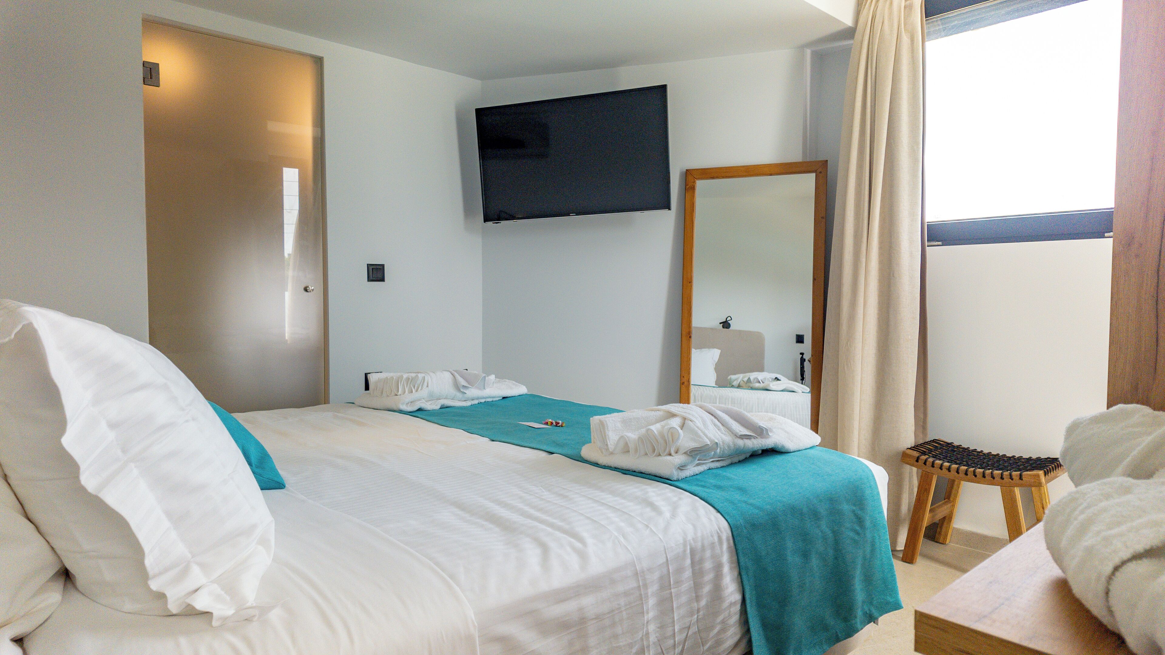 A hotel room with a bed, a television, a mirror, and a small wooden stool.