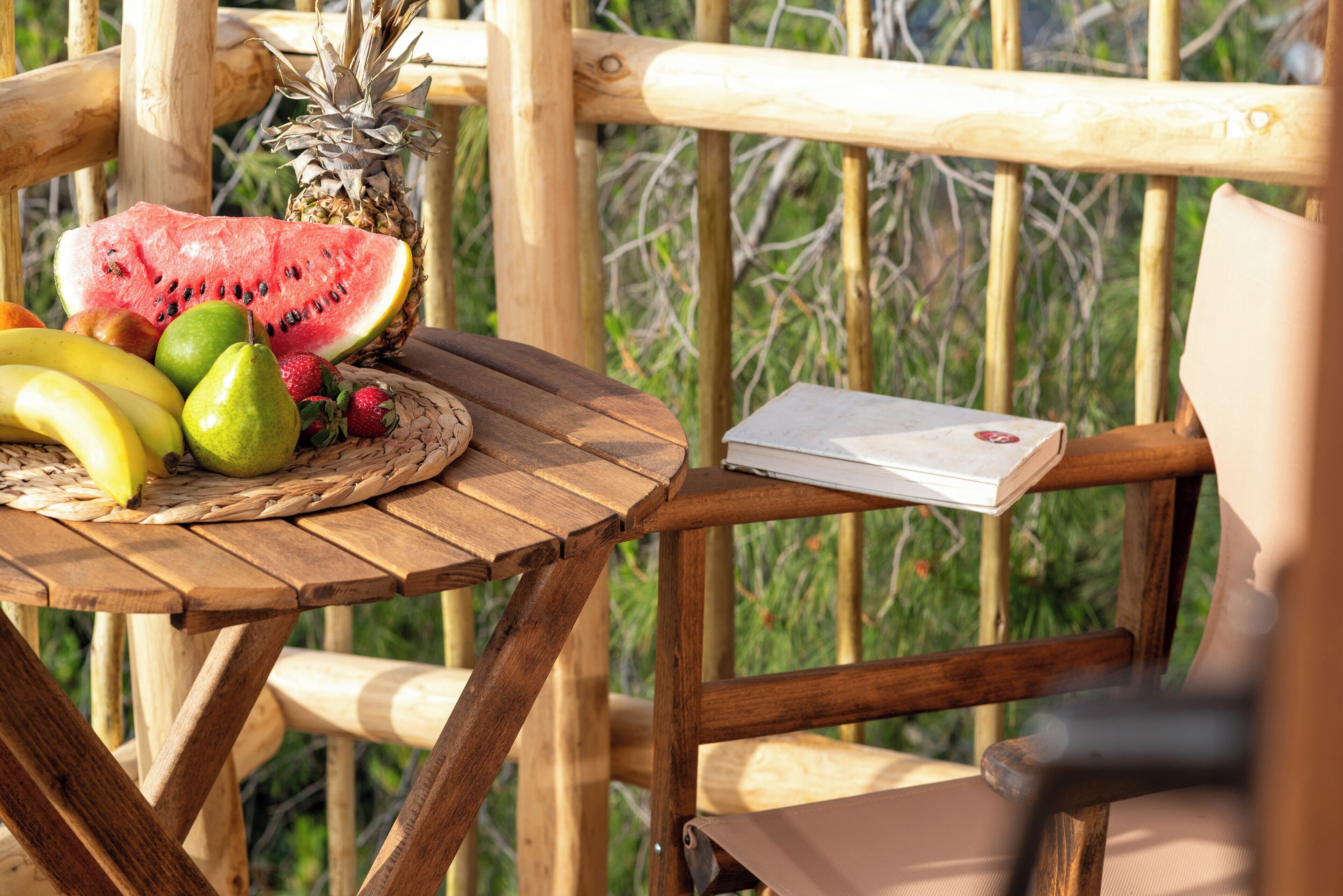 A wooden table with a woven placemat, a pineapple, watermelon, bananas, pears, and strawberries. A book is placed on the table.
