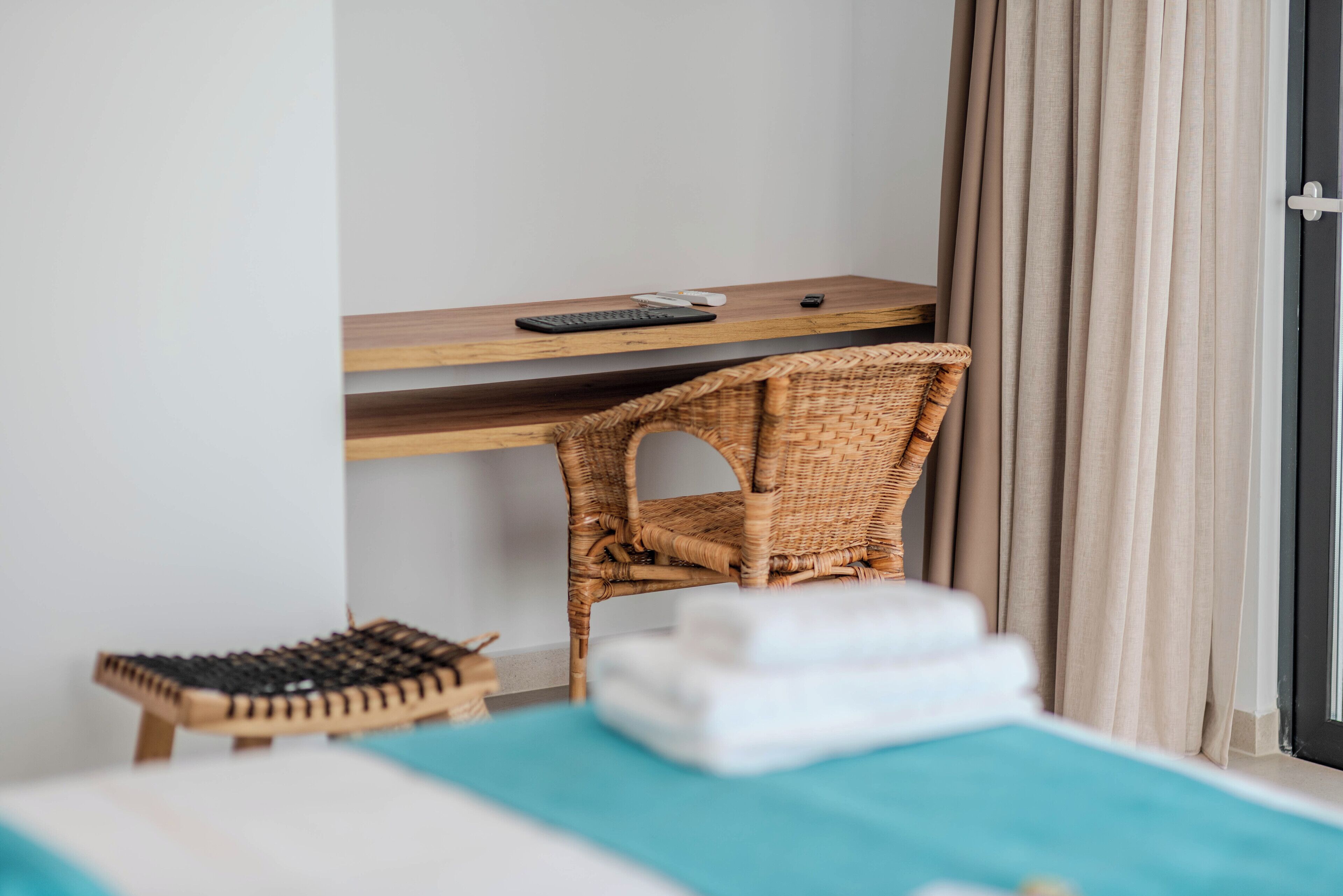 A hotel room with a bed, a small desk with a keyboard and mouse, a wicker chair, and a small stool.