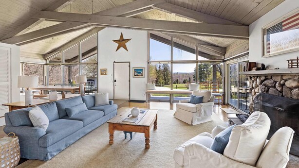 A spacious living room with a high ceiling, a stone fireplace, and large windows offering a view of the outdoors. - Private 5-BR Mountain Estate 10-Min to Manchester (Dorset)