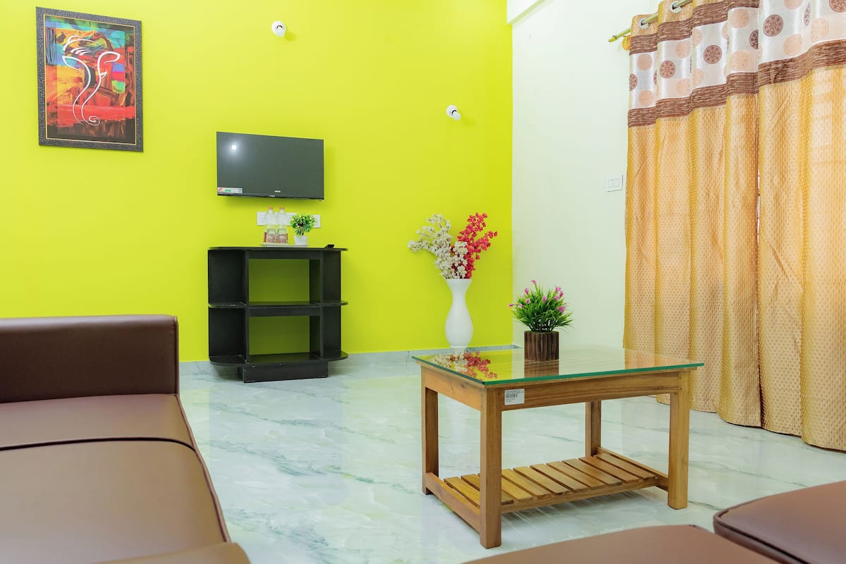 Uma sala de estar com uma parede amarelo vibrante, uma TV de tela plana, uma mesa com tampo de vidro, um sofá marrom e um vaso de planta.