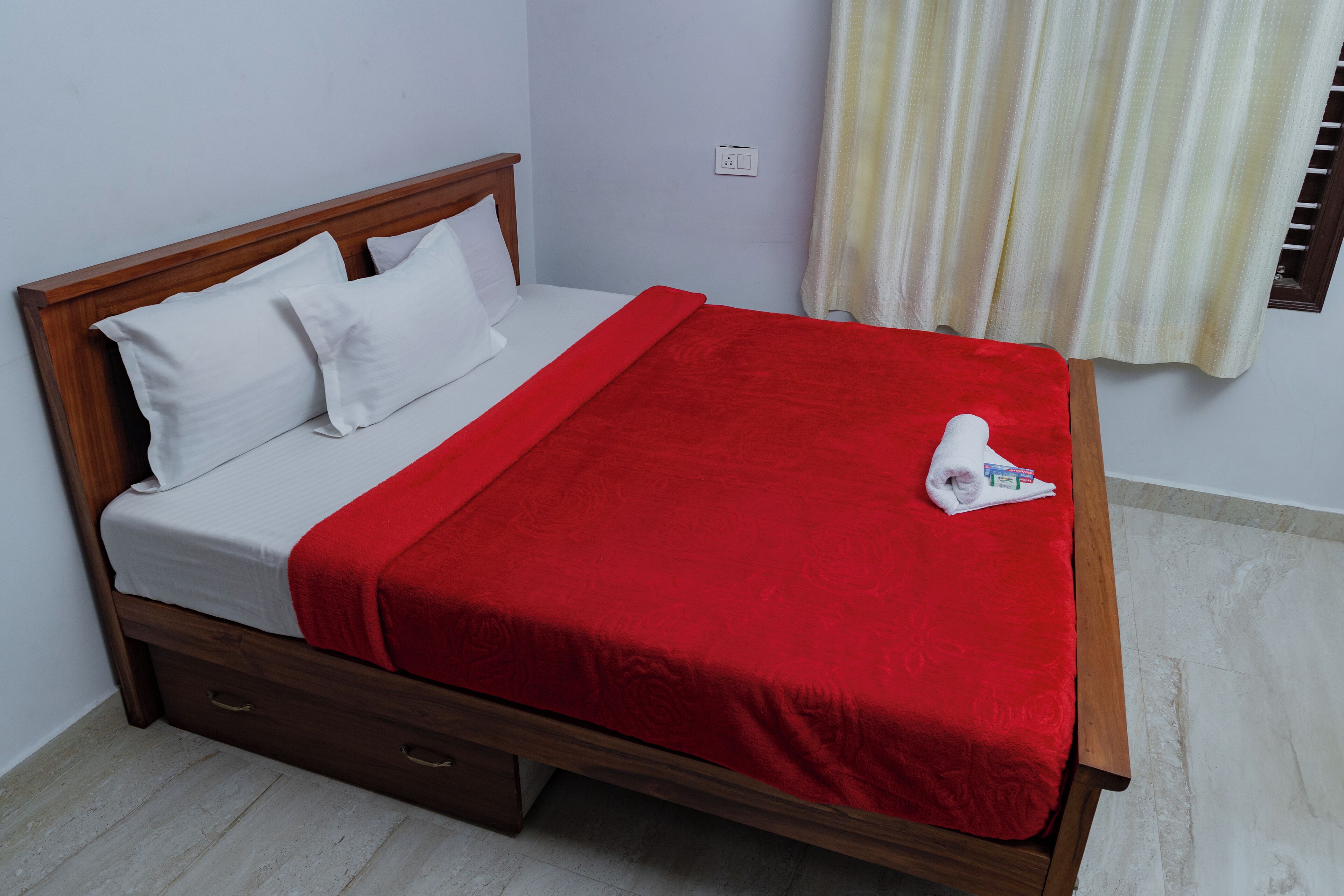 Una cama bien hecha con una manta roja y almohadas blancas, cabecera de madera y un pequeño cajón auxiliar.
