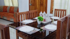 A dining area with a glass table surrounded by wooden chairs, a wooden door, and a sofa with orange cushions. - Coorg Residency Homestay (Somvarpet)