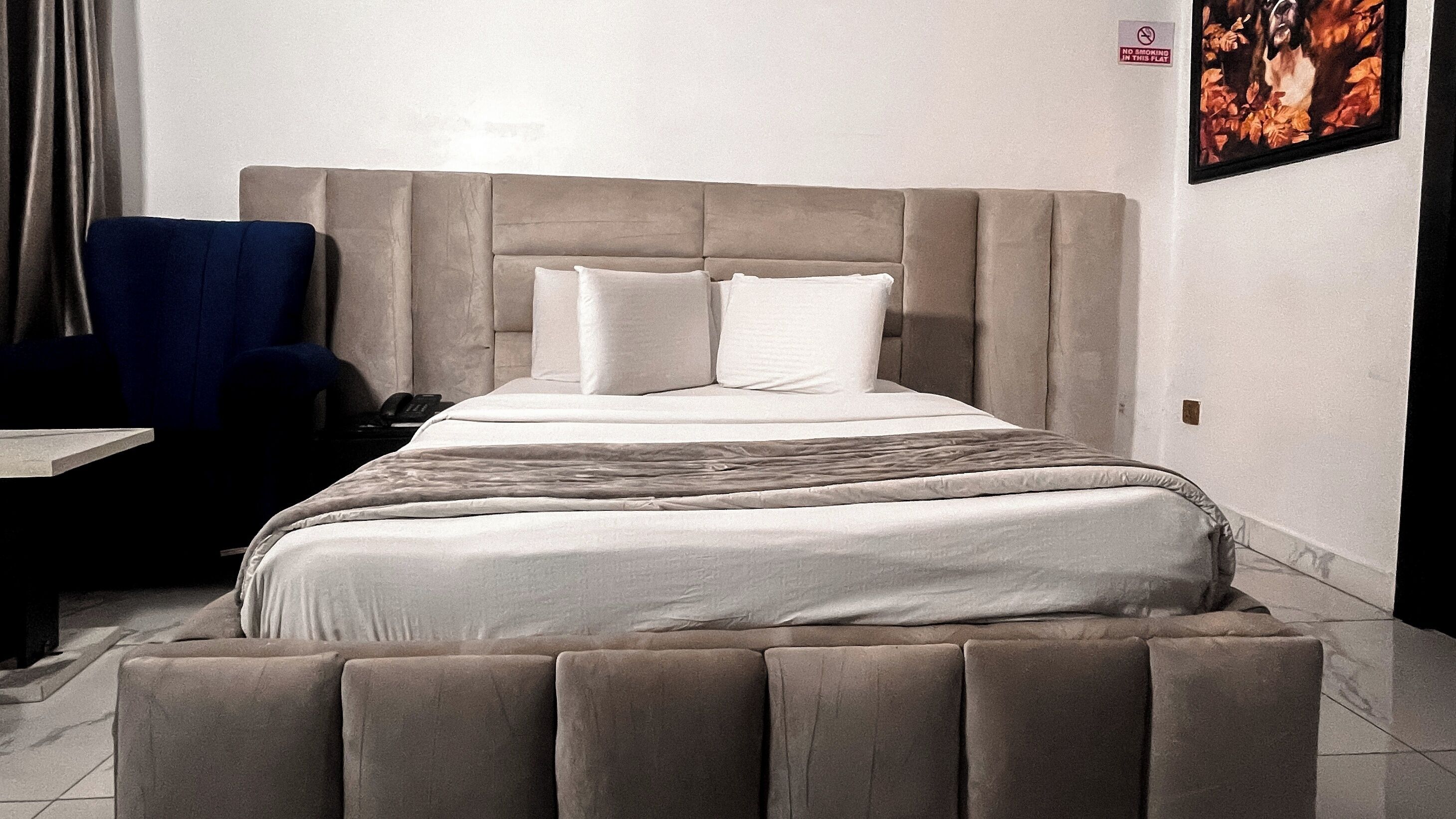 Habitación de hotel con una cama grande, cabecera, mesita de noche, una silla y un cuadro en la pared.