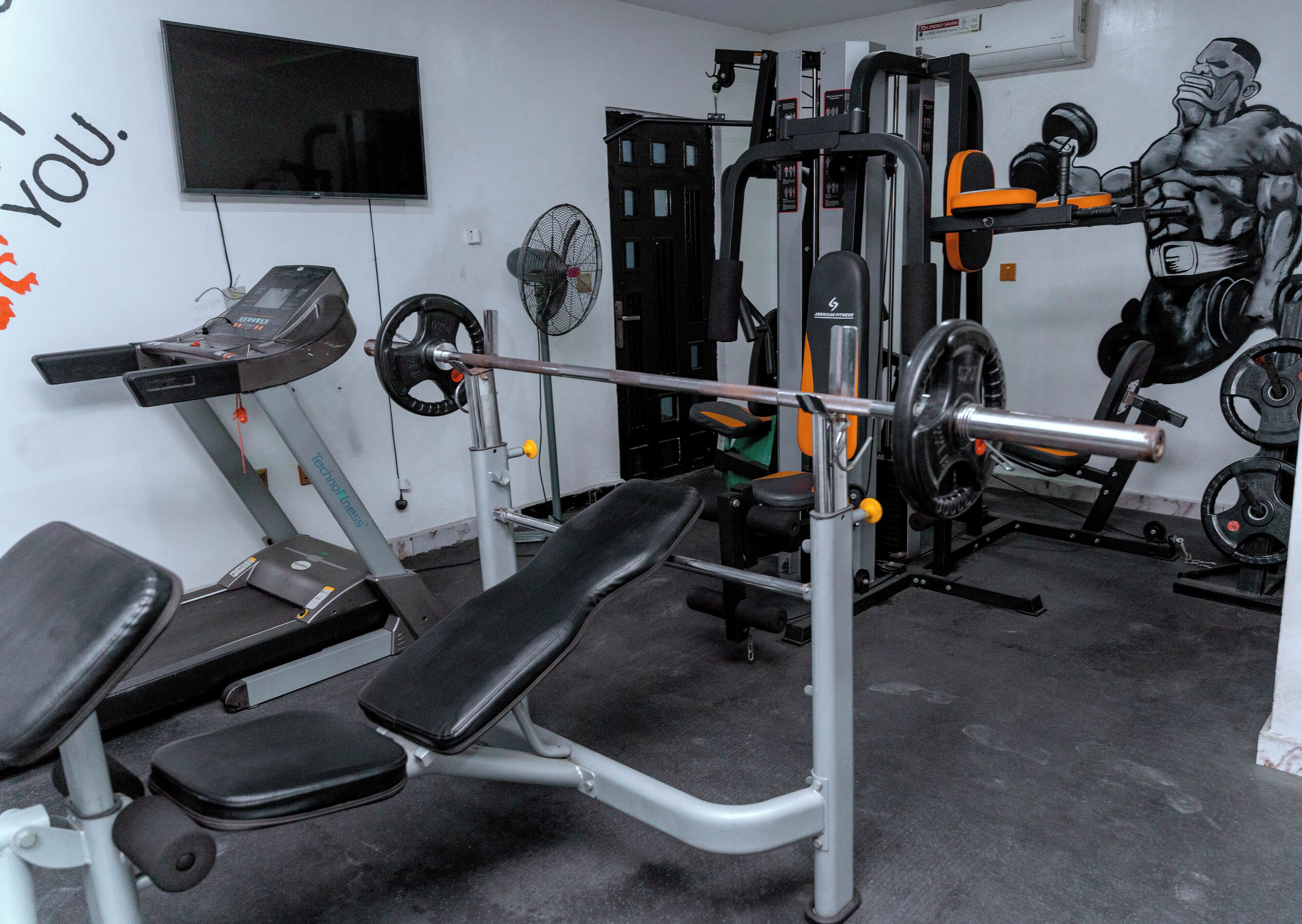Un gimnasio con equipo de levantamiento de pesas, una banca y un televisor montado en la pared.