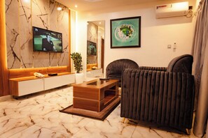 A modern living room with a marble floor, a flat-screen TV mounted on the wall, a dark sofa, a wooden coffee table, and a potted plant. - 2-bedroom Flat Lekki Phase 1 - Pool - Spa (Lekki)
