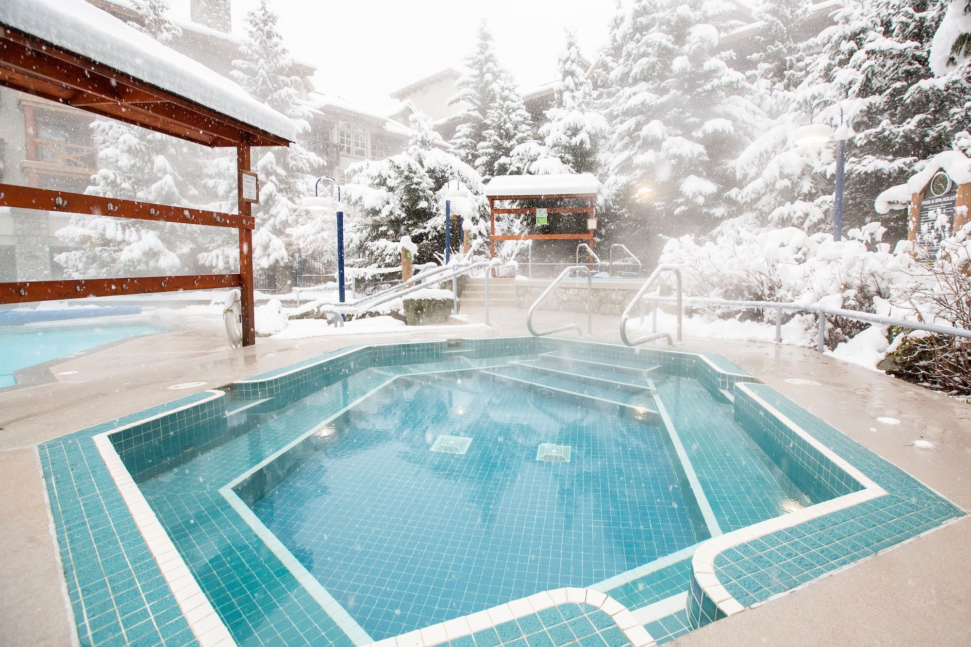 A swimming pool with a tiled deck, surrounded by snow-covered trees and a wooden structure.