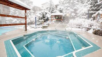 A swimming pool with a tiled deck, surrounded by snow-covered trees and a wooden structure.