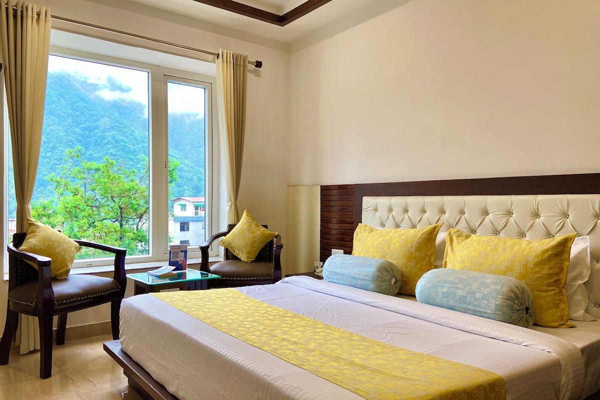 A hotel room with a large bed, a chair, a table, and a view of greenery outside.