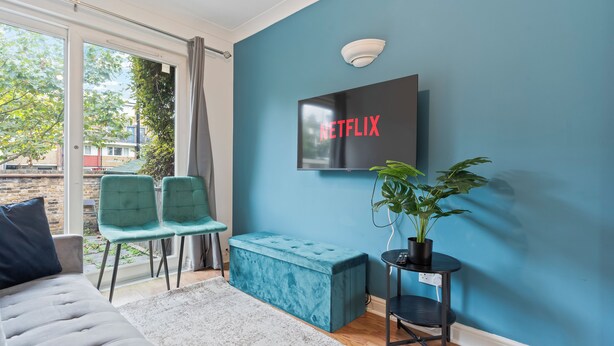 A modern living room with a teal sofa, a grey rug, a blue bench, and a TV mounted on a blue wall displaying "NETFLIX". - Cozy London Townhouse With 4 Bedrooms (London)