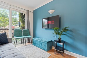 A modern living room with a teal sofa, a grey rug, a blue bench, and a TV mounted on a blue wall displaying "NETFLIX". - Cozy London Townhouse With 4 Bedrooms (London)