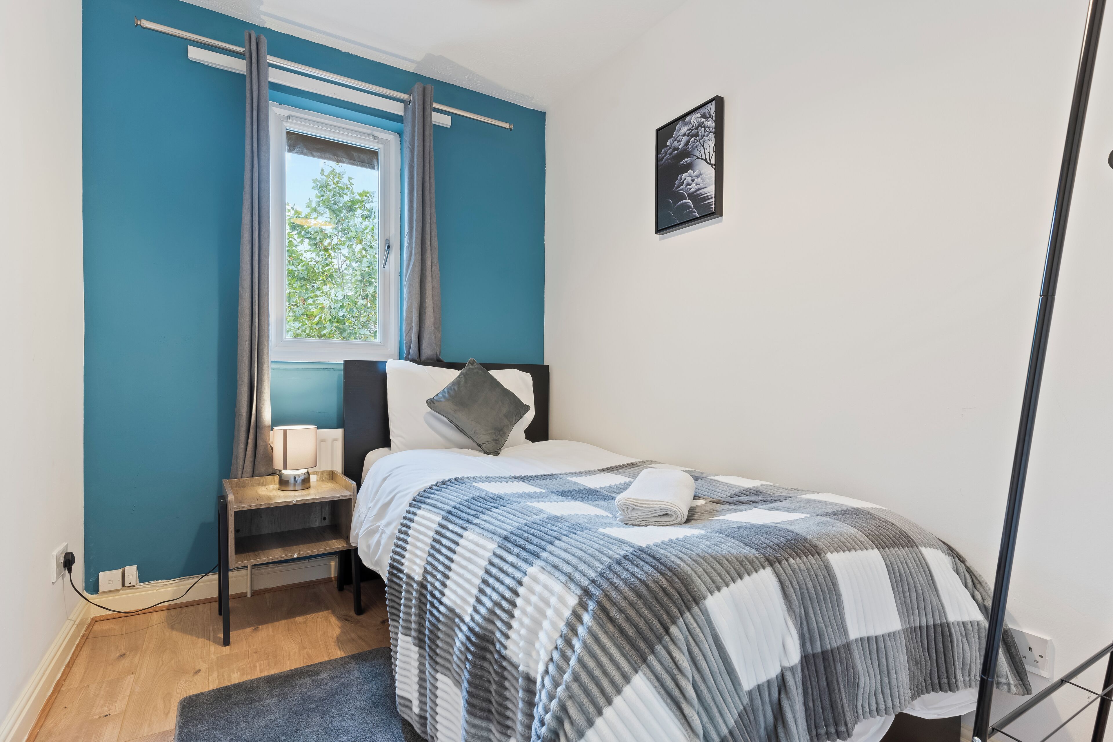 Un dormitorio con cama, mesita de noche, lámpara, ventana con cortinas e imagen colgada en la pared.