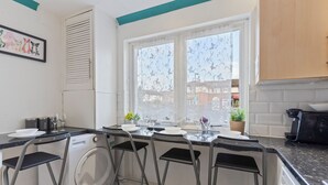 A compact kitchen with a built-in washer, bar stools, a window with patterned curtains, and a view of buildings outside. - Cozy London Townhouse With 4 Bedrooms (London)