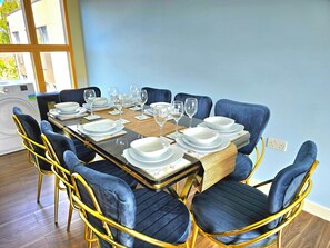 A dining table set with white plates, bowls, and wine glasses, surrounded by blue upholstered chairs with gold frames, in a room with wooden flooring and a large window. - Retreat Private Rooftop 3 Bedroom Apartment (London)