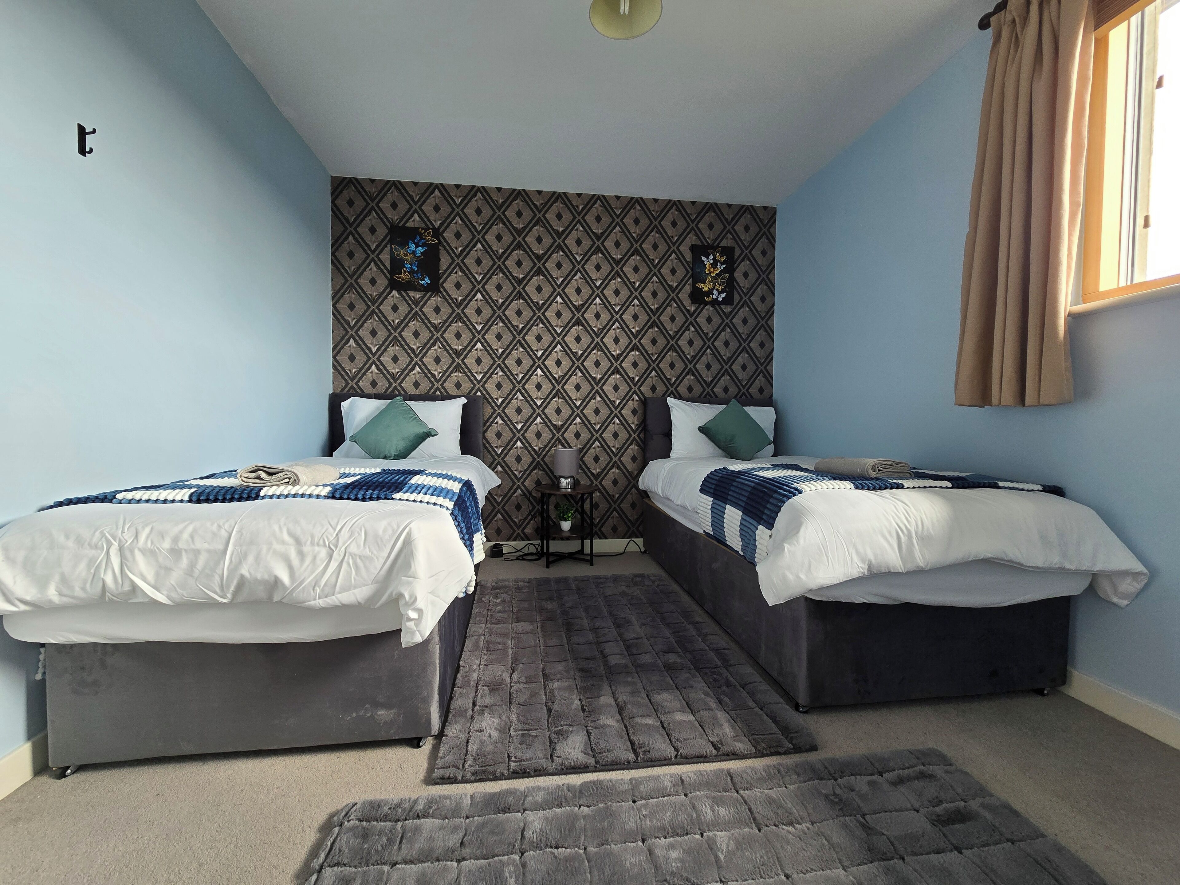 A bedroom with two beds, a patterned wall, a small table with a lamp, and a window with curtains.