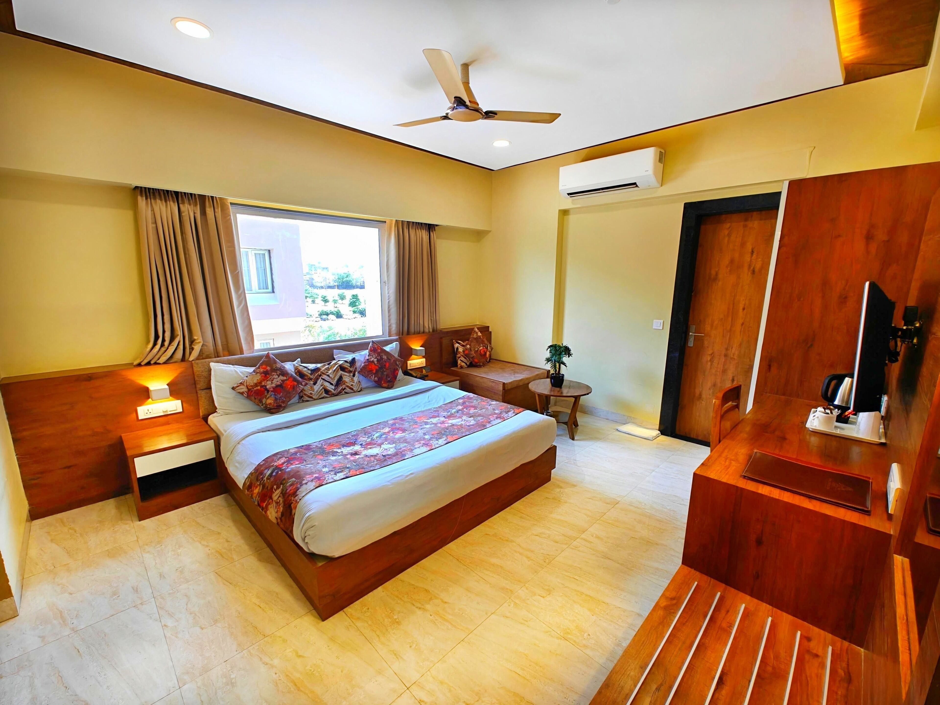 A hotel room with a large bed, bedside tables, a desk, a chair, a ceiling fan, an air conditioning unit, and a window with curtains.